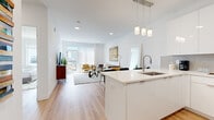 Located at 5711 John F. Kennedy Boulevard in North Bergen, NJ, this contemporary single-floor residence showcases a thoughtfully designed open-concept layout finished with light hardwood flooring and crisp white walls throughout. The modern kitchen is outfitted with high-gloss white cabinetry extending to the ceiling, a subway tile backsplash, stainless steel appliances including a gas range, microwave, and French door refrigerator, and a large white quartz island with pendant lighting — ideal for both cooking and casual dining. The island seamlessly connects to a dining area and a bright, open living room. The living space is anchored by large sliding glass doors that flood the room with natural light and open directly onto a private balcony with outdoor views. The bedroom offers a serene retreat with a wide window framing neighborhood views and warm hardwood flooring. The bathroom features striking blue marble-style tile in the tub surround, white marble-look floor tile, a wood-grain floating vanity with a backlit medicine cabinet, and brushed gold fixtures, creating a refined, spa-like atmosphere. The entry foyer provides a welcoming transition into the home, complete with ample cabinetry for storage. The overall design balances clean, minimalist aesthetics with warm, inviting finishes.