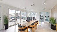 Located at 5832 Coastal Pointe Dr in Virginia Beach, VA, this single-floor community amenity space offers a well-appointed and thoughtfully designed environment for socializing, recreation, and productivity. The layout is divided into distinct functional zones that flow seamlessly into one another. A bright dining and gathering area anchors one end of the space, featuring large-format tile flooring, floor-to-ceiling glass walls, and sliding doors that open directly onto an expansive outdoor terrace with patio seating and sweeping views of the surrounding community. Adjacent to this is a casual lounge area furnished with plush seating, warm-toned carpet tiles, curated artwork, and a wall-mounted TV — ideal for relaxed gatherings or quiet downtime. A dedicated game room provides recreational amenities including ping-pong and billiards tables beneath sleek linear pendant lighting, accented by bold wall art. A co-working or collaborative area is equipped with a large communal table, modern chairs, warm wood-paneled walls, and direct elevator access. Glass-partitioned rooms offer semi-private spaces for focused work or small meetings. Throughout, the design is characterized by clean white walls, abundant natural light, and contemporary furnishings, creating a bright, welcoming, and versatile community hub.