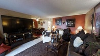 This single-floor residence offers a thoughtfully arranged and richly decorated interior that blends comfort with classic style. The living area serves as the heart of the space, featuring a bold terracotta accent wall adorned with framed wall art and decorative sconces, creating a warm and inviting focal point. Dark wood furniture, including an ornate coffee table and a classic leather sofa, are complemented by plush, decorative pillows and a deep-toned area rug that anchors the seating arrangement. A large flat-screen television is mounted on an entertainment console along one wall, making the room well-suited for both relaxation and entertainment. A white slipcovered armchair adds a contrasting design element, contributing to the layered aesthetic of the room. The space flows naturally into a dining area, visible in the background, which features a dark wood dining table and matching chairs beneath a ceiling fan with light fixture. The textured ceiling and neutral carpet throughout lend a cohesive quality to the interior. Soft table lamps provide ambient lighting, enhancing the cozy atmosphere. Overall, the space presents a well-furnished, single-level home with a consistent design language rooted in warm tones and traditional furnishings.