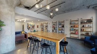 This single-floor communal space blends industrial architecture with contemporary design, creating a dynamic and inviting environment. The exposed concrete ceiling, complete with visible structural beams and conduit, contrasts with polished concrete floors throughout, establishing a raw yet refined aesthetic. Large street-facing windows flood the space with natural light, offering urban views and enhancing the open, airy atmosphere.

The layout is thoughtfully divided into distinct functional zones. A generous communal high-top table crafted from natural wood, surrounded by sleek black bar stools, serves as a central gathering point, illuminated overhead by a sculptural brass-and-glass globe chandelier. Wall-mounted open shelving units display books and decorative objects, adding warmth and visual interest.

Multiple lounge areas are furnished with colorful seating — including emerald green armchairs, blue sofas, and a round white bistro table with teal chairs — layered over patterned area rugs. Bold original artwork and a large-scale painted cityscape mural serve as striking focal points throughout the space. A stainless steel elevator and entry vestibule with tall windows complete the well-appointed layout.