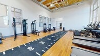 This expansive fitness center offers a generously sized, single-floor layout designed to accommodate a comprehensive range of workout activities. The space is defined by its striking architectural character, featuring soaring ceilings with exposed wood beam construction and visible ductwork that lend an industrial yet inviting atmosphere. Large, arched multi-pane windows line the walls, flooding the interior with abundant natural light and providing views of the surrounding urban brick buildings. The warm hardwood floors extend throughout the space, complemented by a prominent black turf sprint track running centrally down the length of the room — marked with yard-line indicators for performance training. The cardio zone is well-appointed with treadmills, ellipticals, stationary bikes including a Peloton, and rowing machines, all arranged along the windowed walls. A dedicated strength training area features a large functional training rig with cable attachments, an extensive dumbbell rack, adjustable benches, and multiple selectorized weight machines. Full-length mirrors are positioned along one wall to support form monitoring. The layout is open and uncluttered, providing ample space for movement between equipment zones, making this facility suitable for a wide variety of fitness routines.