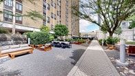 Located at 450 North 18th Street in Philadelphia's vibrant Art Museum neighborhood, this thoughtfully designed outdoor courtyard serves as a communal amenity space nestled between high-rise residential buildings. The grounds are generously landscaped with mature trees, manicured shrubs, and lush garden beds bursting with flowering hydrangeas and dense hedgerows, creating a green retreat from the surrounding urban environment.

The courtyard is organized into distinct activity zones connected by herringbone-patterned brick walkways and gravel pathways. A lounge area features warm teak-framed sofas and armchairs with neutral cushions, arranged around a low coffee table for relaxed outdoor gathering. Adjacent to this is a formal outdoor dining area furnished with sleek dark-framed tables and chairs, well-suited for group meals or social events.

A dedicated grilling station equipped with stainless steel gas grills provides a functional space for outdoor cooking, tucked within a landscaped alcove surrounded by greenery. Adirondack-style wooden chairs are positioned throughout the grounds, offering quiet spots to relax among the trees. A rugged stone retaining wall adds architectural character to one edge of the space, complementing the natural materials used throughout. Bollard lighting is distributed across the courtyard, ensuring the space remains accessible and inviting after dark.