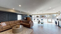 This single-floor space is thoughtfully designed as a modern communal lounge, blending distinct functional zones within an open, flowing layout. The flooring throughout features a rich wood-look herringbone pattern that adds visual depth and warmth. A striking dark navy geometric accent wall serves as a dramatic focal point, anchoring the lounge and media area, where a large sectional sofa, a round coffee table, and a flat-screen television create an inviting, relaxed setting. Vibrant abstract artwork on the wall adds a bold pop of color to this zone.

Toward the front entrance, a well-appointed beverage bar showcases navy cabinetry, a light stone countertop, under-counter refrigeration, and an eye-catching black-and-white patterned tile backsplash, framed by warm pendant lighting. Glass double doors at the entry provide a welcoming and bright transition from the exterior.

The opposite end of the space opens expansively toward floor-to-ceiling windows and glass doors, flooding the interior with abundant natural light and offering direct views of an outdoor court. This area features bar-height seating, clustered ottomans, and large bronze dome pendant lights suspended overhead, creating a casual and social atmosphere ideal for gathering.