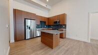 This well-designed single-floor unit showcases a clean, contemporary aesthetic throughout. The open-concept kitchen serves as the heart of the space, featuring rich warm-toned wood cabinetry that extends to impressive heights, complemented by a striking teal subway tile backsplash. A central kitchen island with a light grey countertop provides additional workspace and casual seating potential. Stainless steel appliances, including a French door refrigerator with an ice and water dispenser, a built-in microwave, dishwasher, and range, complete the modern kitchen setup. Light wood-style flooring flows through the kitchen and living areas, enhancing the bright, airy feel created by the soaring ceilings. The living area benefits from abundant natural light through a large window and direct access via a glass-panel door. The bedrooms feature plush neutral carpeting, generous windows with blinds, and tall ceilings that lend each room an open, comfortable atmosphere. The bathrooms are finished with the same warm wood cabinetry found in the kitchen, paired with white countertops, large vanity mirrors, and light wood-style flooring. One bathroom includes a glass-enclosed walk-in shower with built-in shelving, while another features a clean, functional layout. Throughout, the neutral white walls and consistent design language create a cohesive and inviting living environment.