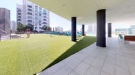 This contemporary amenity level showcases a thoughtfully designed outdoor and semi-covered retreat set against a striking urban skyline. The space is unified by large-format light grey tile flooring throughout, complemented by grey walls and smooth concrete columns that anchor the covered zones with a sleek, modern aesthetic.

A spacious social lounge area features multiple round dining tables paired with vibrant yellow and orange chairs, a wall-mounted flat-screen display, and tall tropical potted plants that bring warmth and greenery indoors. This gathering zone connects seamlessly to an open terrace furnished with colorful woven accent chairs beside a glass balustrade, offering open views of the surrounding cityscape.

A standout feature is the expansive artificial turf lawn, ideal for recreation and relaxation, lined with blue lounge chairs overlooking what appears to be a pool area. Bold cylindrical dark columns frame this outdoor zone, creating a resort-like atmosphere flooded with natural light.

Additional areas include a bar counter clad in decorative geometric-patterned tile, a utility corridor with clean grey walls and architectural plant accents, and a grand staircase leading between levels. Lush landscaped planters with tropical foliage add a natural softness throughout, making this a well-rounded, amenity-rich communal space designed for both active use and leisurely enjoyment.