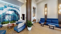 This single-floor lobby space showcases a thoughtfully designed interior that balances modern aesthetics with functional comfort. Soaring double-height ceilings lend an expansive, airy quality to the space, amplified by large arched glass entry doors and strategically placed windows that frame views of lush outdoor greenery, including mature palm trees.

The main seating area is anchored by generous L-shaped and curved blue upholstered sofas, complemented by sculptural white lounge chairs and low round side tables. Warm walnut wood accents appear throughout—on vertical slat columns, floating vanity cabinetry, and integrated sofa consoles—providing a rich contrast to the crisp white walls and light-toned tile flooring.

A standout feature is the large-scale abstract wall mural rendered in vivid blues and teals, adding a dramatic focal point. Unique looped sculptural sconces mounted on the wood columns deliver warm ambient lighting. A floating walnut vanity with hexagonal white tile backsplash and a framed mirror adds a refined finishing touch near the entry corridor. Potted tropical greenery is placed throughout, enhancing the space's fresh, welcoming ambiance.