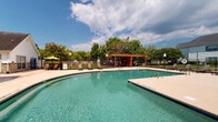 Located at 1530 Wimbledon Dr in Greenville, North Carolina, this outdoor community amenity space centers around a generously sized swimming pool with clear, blue-green water. The pool area is designed for both relaxation and recreation, featuring multiple entry points with stainless steel handrails and a shallow end clearly marked for safety. Surrounding the pool is a broad concrete deck outfitted with lounge chairs and chaise seating, providing ample space for sunbathing and outdoor gatherings. A wooden pergola with an attached shade structure anchors one side of the deck, offering a shaded retreat with additional seating beneath. A colorful patio umbrella adds a pop of character near an outdoor dining table and chairs. The clubhouse building, constructed with warm natural stone accents and a pitched roof with a chimney, borders the pool area and serves as an architectural focal point. The property is fully enclosed by a black iron fence, ensuring privacy and security. Lush mature trees line the perimeter, providing a green backdrop and natural shade. Views of the broader residential community, including neighboring multi-story homes, are visible beyond the fence, reinforcing the sense of a well-appointed neighborhood amenity designed for community enjoyment.