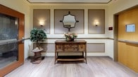 Located at 130 Rollins Ave in Rockville, Maryland, this elevator lobby presents a refined and welcoming atmosphere designed with classic elegance in mind. The single-floor space is finished with light wood-look flooring that runs throughout, providing a clean and bright foundation for the overall design.

The walls are adorned with decorative framed paneling accented in warm gold tones, set against a soft cream and taupe color palette that lends a sophisticated, timeless character to the space. Recessed lighting and wall sconces provide warm, layered illumination, enhancing the inviting ambiance of the lobby.

A standout decorative vignette features a rich, dark-wood console table topped with a floral arrangement, flanked by an ornate wall mirror and wall sconces — adding a boutique-hotel quality to the arrival experience. A potted indoor tree positioned nearby introduces a touch of natural greenery to the space.

Dual elevator doors finished in a neutral tone offer convenient vertical access, while a set of warm wood-framed double doors with glass panels provides a clearly defined entry and exit point. Safety and wayfinding elements, including exit signage and fire safety equipment, are thoughtfully integrated without detracting from the overall aesthetic. The lobby successfully balances functional building circulation with an upscale, hospitable design.