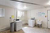 Located at 3901 Hammerberg Rd in Flint, Michigan, this single-floor laundry and utility room provides a functional, shared-use space designed to serve a multi-unit building. The room is finished with white-painted walls and a flat ceiling with flush-mounted lighting, keeping the interior bright and practical.

The laundry area is equipped with commercial-grade top-load washers and a front-loading dryer, accompanied by a freestanding utility sink that offers added convenience for hand-washing or cleaning tasks. Dryer exhaust ducting is routed through the space and vented to the exterior, and a baseboard heating unit runs along the wall beneath a window fitted with vertical blinds, ensuring year-round usability. Instructional signage is posted on the walls to guide users through the laundry process.

A dedicated utility area within the room houses the building's mechanical infrastructure, including a central heating unit, a large water heater, and an extensive network of overhead pipes and conduit. A large multi-meter electrical panel serves the individual units of the building and is mounted prominently on one wall, alongside a fire extinguisher for safety. A full-size refrigerator and an electrical panel box are also present. The concrete floor and exposed utility components reflect the room's practical, no-frills character, while a solid-panel entry door provides secured access to the space.