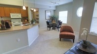 This single-story residence showcases an open-concept layout that connects the kitchen, dining, and living areas into one cohesive, flowing space. The kitchen is positioned along one side, featuring warm wood cabinetry, a countertop breakfast bar with pendant lighting overhead, a built-in sink, and an adjacent built-in shelving unit that adds both storage and visual interest. Track lighting runs along the kitchen ceiling, providing focused illumination across the workspace.

The dining area sits centrally within the open floor plan, accommodating a glass-top table surrounded by dark-framed chairs, positioned near a bright window dressed with horizontal blinds that allow natural light to filter in. Neutral carpeting extends throughout the living and dining zones, contributing to the warm, understated ambiance of the space.

Crown molding trims the upper edges of the walls, adding a refined architectural detail that elevates the overall aesthetic. The open sightlines between each zone create a sense of spaciousness, making the layout feel connected and functional. The muted green-gray wall tones complement the warm furnishings and flooring, resulting in a cohesive, comfortable interior environment.