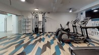 This fitness center is a thoughtfully designed, single-floor workout facility that combines functional equipment with modern aesthetics. The most striking design element is the bold chevron-patterned flooring, featuring a mix of light wood, charcoal, and vibrant blue tones that energize the entire space and set a dynamic, motivational atmosphere.

The cardio zone is anchored by a row of Life Fitness treadmills and elliptical machines, positioned in front of a statement accent wall adorned with fitness-themed typography — featuring words like "Strength," "Fitness," "Flexibility," and "Nutrition" — reinforcing an active and goal-oriented environment. Flat-screen monitors are mounted for entertainment during workouts.

The strength training area is well-appointed with a multi-functional cable machine, adjustable weight benches, and a full dumbbell rack with a large mirror wall that visually expands the space and supports proper form. Additional open shelving provides storage for accessories and equipment.

The facility is illuminated by recessed and pendant lighting, keeping the space bright and welcoming. A glass-door entry connects seamlessly to an adjacent common area, offering an open and accessible feel. Overall, this fitness center delivers a complete workout experience in a clean, modern, and visually engaging environment.