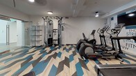 This fitness center is a thoughtfully designed, single-floor workout facility that combines functional equipment with modern aesthetics. The most striking design element is the bold chevron-patterned flooring, featuring a mix of light wood, charcoal, and vibrant blue tones that energize the entire space and set a dynamic, motivational atmosphere.

The cardio zone is anchored by a row of Life Fitness treadmills and elliptical machines, positioned in front of a statement accent wall adorned with fitness-themed typography — featuring words like "Strength," "Fitness," "Flexibility," and "Nutrition" — reinforcing an active and goal-oriented environment. Flat-screen monitors are mounted for entertainment during workouts.

The strength training area is well-appointed with a multi-functional cable machine, adjustable weight benches, and a full dumbbell rack with a large mirror wall that visually expands the space and supports proper form. Additional open shelving provides storage for accessories and equipment.

The facility is illuminated by recessed and pendant lighting, keeping the space bright and welcoming. A glass-door entry connects seamlessly to an adjacent common area, offering an open and accessible feel. Overall, this fitness center delivers a complete workout experience in a clean, modern, and visually engaging environment.