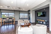 This thoughtfully designed single-floor commercial space offers a blend of functionality and contemporary style across several distinct zones. The main lounge area serves as a welcoming centerpiece, featuring plush, curved cream upholstered seating arranged around a warm wood coffee table atop a woven area rug. A recessed dark-accent niche provides a focused wall-mounted display, creating an inviting media corner ideal for gatherings or presentations. Large industrial-style overhead garage windows flood the space with natural light and offer views of the surrounding cityscape, lending an open, airy feel to the interior. Rich hardwood flooring runs throughout the main areas, grounding the neutral gray walls with warmth and texture. A high-top dining table with woven-seat bar stools provides a casual dining or collaboration spot bathed in natural light from nearby windows. A secondary lounge area offers additional seating with curated wall art and soft globe sconces, making it well-suited for quieter, more intimate settings. The breakroom kitchen is practical and well-appointed, featuring light wood cabinetry, ample counter space, a full-size refrigerator, and a bold red floor that adds a vibrant pop of color. A central island table offers additional workspace or casual dining. The drop-panel ceiling with recessed and fluorescent lighting maintains a clean, professional atmosphere throughout the entire space.