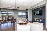 This thoughtfully designed single-floor commercial space offers a blend of functionality and contemporary style across several distinct zones. The main lounge area serves as a welcoming centerpiece, featuring plush, curved cream upholstered seating arranged around a warm wood coffee table atop a woven area rug. A recessed dark-accent niche provides a focused wall-mounted display, creating an inviting media corner ideal for gatherings or presentations. Large industrial-style overhead garage windows flood the space with natural light and offer views of the surrounding cityscape, lending an open, airy feel to the interior. Rich hardwood flooring runs throughout the main areas, grounding the neutral gray walls with warmth and texture. A high-top dining table with woven-seat bar stools provides a casual dining or collaboration spot bathed in natural light from nearby windows. A secondary lounge area offers additional seating with curated wall art and soft globe sconces, making it well-suited for quieter, more intimate settings. The breakroom kitchen is practical and well-appointed, featuring light wood cabinetry, ample counter space, a full-size refrigerator, and a bold red floor that adds a vibrant pop of color. A central island table offers additional workspace or casual dining. The drop-panel ceiling with recessed and fluorescent lighting maintains a clean, professional atmosphere throughout the entire space.