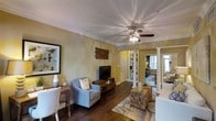 This single-floor residence showcases a thoughtfully designed interior with a warm, cohesive aesthetic throughout. The main living area features rich, dark hardwood flooring that anchors the space, complemented by soft yellow-toned walls that lend a welcoming ambiance. Crown molding lines the ceiling, adding a refined architectural detail, while a ceiling fan with an integrated light fixture serves as both a functional and decorative element overhead.

The living room is well-appointed, with space for a comfortable seating arrangement, a media console housing a flat-screen television, and accent furniture including rustic wooden side tables that add textural contrast. A framed wall piece and a natural wood wall accent contribute to the room's layered, curated décor.

A distinctive white decorative column marks the transition between the living area and an adjacent space, creating a sense of separation while maintaining an open feel. Built-in shelving is visible beyond the column, offering both storage and display potential. Additional rooms are accessible through the connected layout, suggesting a well-organized and functional floor plan. Natural light filters in from windows in the adjoining rooms, brightening the overall interior.