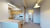 This single-floor space offers a functional and comfortable layout designed for everyday living. The kitchen is a standout feature, boasting light wood cabinetry that extends to the ceiling, providing ample storage. It is equipped with a white electric range, an under-cabinet range hood, and a sink set within a continuous countertop. A kitchen island adds extra prep space and separates the cooking area from the rest of the living space. A warm pendant light hangs overhead, casting a welcoming glow over the area. Exposed ceiling beams add a subtle architectural character to the kitchen and adjoining spaces. The flooring transitions from hard surface in the kitchen to plush, neutral-toned carpet in the open living area, contributing to a cozy atmosphere throughout. A hallway visible from the main living area leads to additional rooms, with a window at the far end drawing in natural light and enhancing the sense of depth. The overall color palette is soft and neutral, with light gray and beige tones throughout the walls, ceilings, and floors, creating a calm, cohesive environment. The textured ceiling adds a layer of visual interest and complements the understated, practical design of the space.