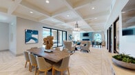This sophisticated club room offers a thoughtfully designed, open-plan layout that seamlessly blends social, dining, and recreational spaces. The interior is anchored by light-toned herringbone-patterned flooring that flows throughout, complemented by elegant coffered ceilings with recessed lighting and pendant fixtures that create a bright, airy ambiance.

The main gathering area features a substantial rustic wood dining table surrounded by upholstered chairs, alongside a comfortable lounge seating arrangement centered around a sleek fireplace clad in teal glass tile. A wall-mounted television above the fireplace and an arched window offering outdoor views complete this inviting focal point.

The full demonstration kitchen is a standout feature, showcasing a large waterfall-edge quartz island with bar seating, a gas cooktop, stainless steel appliances, and a striking mosaic glass tile backsplash in earthy teal and green tones. Open wood shelving adds a warm, functional touch.

A dedicated billiards room provides recreational space, outfitted with a full-size pool table and arched windows that flood the room with natural light and views of palm-lined outdoor areas. Intimate seating nooks with arched windows offer quiet spaces for relaxed conversation, while glass-paneled walls maintain visual connectivity throughout the entire club room.