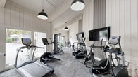 Located at 7910 Crossroads Dr in North Charleston, SC, this fitness center is a well-appointed, single-floor exercise facility designed to support a full range of workout routines. The space features soaring vaulted ceilings with exposed white-painted beams and shiplap wall paneling that lend a clean, modern-farmhouse aesthetic throughout.

Natural light floods the interior through expansive floor-to-ceiling windows and glass doors, offering views of the surrounding outdoor area, including a pool patio visible from the main workout floor. Stylish black pendant lights hang from the pitched ceiling, providing warm supplemental lighting. High clerestory windows along the upper walls further enhance brightness and airiness.

The exercise room is outfitted with a comprehensive selection of TRUE-brand cardio equipment, including treadmills, elliptical trainers, and stationary bikes, all positioned facing wall-mounted flat-screen televisions. A multi-functional cable machine, free weights, stability balls, and adjustable workout benches round out the strength and flexibility offerings. The flooring consists of durable, textured carpet tiles suited for high-activity use.

An exposed brick accent wall adds architectural character, while glass-panel doors provide convenient access to the outdoors. The overall ambiance is bright, open, and inviting, creating a motivating environment for exercise.