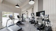 Located at 7910 Crossroads Dr in North Charleston, SC, this fitness center is a well-appointed, single-floor exercise facility designed to support a full range of workout routines. The space features soaring vaulted ceilings with exposed white-painted beams and shiplap wall paneling that lend a clean, modern-farmhouse aesthetic throughout.

Natural light floods the interior through expansive floor-to-ceiling windows and glass doors, offering views of the surrounding outdoor area, including a pool patio visible from the main workout floor. Stylish black pendant lights hang from the pitched ceiling, providing warm supplemental lighting. High clerestory windows along the upper walls further enhance brightness and airiness.

The exercise room is outfitted with a comprehensive selection of TRUE-brand cardio equipment, including treadmills, elliptical trainers, and stationary bikes, all positioned facing wall-mounted flat-screen televisions. A multi-functional cable machine, free weights, stability balls, and adjustable workout benches round out the strength and flexibility offerings. The flooring consists of durable, textured carpet tiles suited for high-activity use.

An exposed brick accent wall adds architectural character, while glass-panel doors provide convenient access to the outdoors. The overall ambiance is bright, open, and inviting, creating a motivating environment for exercise.