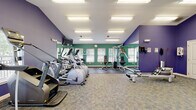 Located at 1320 N 129th Terr in Kansas City, KS, this fitness center is a comprehensive, single-floor facility designed to support a full range of workout routines. The space is immediately distinguished by its bold interior color scheme, featuring vibrant teal and deep purple accent walls that create an energetic atmosphere conducive to exercise.

The cardio section is generously outfitted with multiple treadmills, elliptical trainers, stationary bikes — including both upright and recumbent models — and a WaterRower rowing machine, providing diverse options for cardiovascular training. A multi-station cable and weight machine anchors the strength training area, complemented by a rack of free weights and dumbbells that line the mirrored wall.

Floor-to-ceiling mirrors along one wall visually expand the space and allow users to monitor their form during workouts. The vaulted ceiling adds a sense of openness and volume to the room. Multiple windows fitted with white plantation shutters admit natural light throughout the facility, enhancing the bright, airy feel supported by recessed fluorescent panel lighting overhead.

Flat-screen televisions are mounted at multiple vantage points, offering entertainment during workouts. The flooring transitions between durable carpet tile in the cardio zone and rubber flooring near the weight equipment, balancing comfort and functionality throughout this well-appointed fitness space.