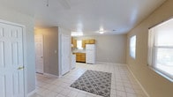 This single-floor space offers an open-concept design that seamlessly connects the living area and kitchen, creating a functional and cohesive layout. The flooring throughout is consistent light-toned ceramic tile, providing a clean, uniform look, complemented by a gray geometric-patterned area rug that anchors the living space.

The kitchen is positioned along one wall and features warm honey-toned wood cabinetry offering ample storage. A white refrigerator, a single-basin sink beneath a window, and under-cabinet space complete the kitchen setup. A white appliance, likely a washer or stove, is visible near a doorway, suggesting additional utility access nearby.

The living area is generously proportioned, benefiting from natural light streaming in through windows on multiple walls. A flush-mount ceiling light fixture provides overhead illumination, and a textured popcorn ceiling adds a classic residential character throughout.

White interior doors and crisp white baseboards contrast neatly against the neutral beige-gray walls, contributing to a bright, tidy atmosphere. The layout is practical and straightforward, making efficient use of the available floor space.