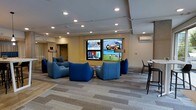 This contemporary lounge is thoughtfully designed to offer a comfortable and engaging environment for relaxation and social interaction. The centerpiece of the room is a striking multi-screen media wall, displaying multiple feeds simultaneously, making it an ideal setting for watching news, sports, and entertainment content. Surrounding the media wall is a carefully curated arrangement of bold blue modular lounge chairs, providing ample seating for groups while adding a vibrant pop of color to the neutral palette of the space. The flooring features a patterned carpet in muted gray tones, contributing to the room's modern aesthetic while adding warmth underfoot. Recessed lighting throughout the ceiling ensures even, ambient illumination across the entire space. A wall-mounted HVAC unit provides climate control, while a high-top table with bar-style seating along the perimeter offers an alternative setting for those who prefer a more upright arrangement. A transitional corridor visible at the far end suggests easy connectivity to adjacent areas. Overall, the space balances a clean, professional design with inviting, comfortable furnishings that encourage both focused viewing and relaxed conversation.
