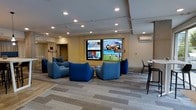 This contemporary lounge is thoughtfully designed to offer a comfortable and engaging environment for relaxation and social interaction. The centerpiece of the room is a striking multi-screen media wall, displaying multiple feeds simultaneously, making it an ideal setting for watching news, sports, and entertainment content. Surrounding the media wall is a carefully curated arrangement of bold blue modular lounge chairs, providing ample seating for groups while adding a vibrant pop of color to the neutral palette of the space. The flooring features a patterned carpet in muted gray tones, contributing to the room's modern aesthetic while adding warmth underfoot. Recessed lighting throughout the ceiling ensures even, ambient illumination across the entire space. A wall-mounted HVAC unit provides climate control, while a high-top table with bar-style seating along the perimeter offers an alternative setting for those who prefer a more upright arrangement. A transitional corridor visible at the far end suggests easy connectivity to adjacent areas. Overall, the space balances a clean, professional design with inviting, comfortable furnishings that encourage both focused viewing and relaxed conversation.