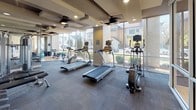 This fitness center is thoughtfully designed to support a comprehensive workout experience in a bright, modern setting. The space is anchored by a generous cardio zone outfitted with treadmills, ellipticals, and stationary bikes, all positioned to take full advantage of the expansive floor-to-ceiling windows that line the perimeter walls. These large glass panels bathe the space in natural light and offer open views of the surrounding neighborhood, creating an energizing atmosphere during any workout. The coffered ceiling adds architectural character overhead, while ceiling fans promote airflow throughout the room. Recessed lighting ensures the space remains well-lit at all hours. A dedicated strength training area features a cable machine and a free weight rack with a full-length mirrored wall, allowing users to monitor form and technique. Wall-mounted televisions are positioned throughout the fitness floor for entertainment during cardio sessions. A separate stretching and flexibility area features hardwood-style flooring, providing a comfortable surface for floor exercises and yoga. The neutral color palette — combining cool gray carpet, warm wood tones, and muted wall hues — lends the center a clean, professional aesthetic that is both functional and visually inviting.