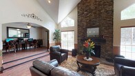 This single-floor home makes a bold first impression with its soaring vaulted ceilings that rise dramatically in the main living area, creating an expansive, airy atmosphere. A floor-to-ceiling stacked stone fireplace serves as the room's focal point, adding texture and warmth with its rich, earthy tones. Above the fireplace, a framed mirror reflects the natural light that pours in generously through large windows, keeping the space bright and inviting throughout the day. The living room is thoughtfully appointed with dark leather seating arranged around a classic round wood coffee table, all grounded by a decorative area rug laid over warm hardwood-style flooring. A tall indoor plant adds a touch of greenery, complementing the natural elements found throughout the design. An elegant arched doorway leads seamlessly into an adjacent room, hinting at the home's well-connected layout and architectural character. The textured ceiling and warm wall tones contribute to a cozy yet refined ambiance. Overall, the space balances casual comfort with distinctive design details, making it both functional and visually compelling for everyday living and entertaining.