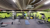 This fitness center in Philadelphia, PA offers a well-equipped, single-floor exercise space designed to support a wide range of workout routines. The room is defined by its bold lime-green accent walls, exposed concrete ceiling, and industrial-style ductwork and piping overhead, giving the space a functional, urban aesthetic. Rubber flooring throughout provides a durable, slip-resistant surface suitable for all types of training.

The cardio section includes treadmills and stationary bikes, complemented by wall-mounted flat-screen monitors for entertainment during workouts. The free weights area features a full dumbbell rack with an extensive range of weights, along with benches and barbells for strength training. A cable machine and additional strength stations are also present to support resistance-based exercises.

Functional training is well-represented with stability balls, medicine balls, resistance bands, foam rollers, and exercise mats organized on dedicated racks. Full-length mirrors line portions of the walls, enhancing the sense of space and allowing users to monitor their form. The facility is climate-controlled with ceiling-mounted HVAC units and is well-lit with overhead fluorescent lighting. A water station is conveniently available within the space. Access is provided through a standard door with clearly marked emergency exits.