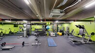 This fitness center in Philadelphia, PA offers a well-equipped, single-floor exercise space designed to support a wide range of workout routines. The room is defined by its bold lime-green accent walls, exposed concrete ceiling, and industrial-style ductwork and piping overhead, giving the space a functional, urban aesthetic. Rubber flooring throughout provides a durable, slip-resistant surface suitable for all types of training.

The cardio section includes treadmills and stationary bikes, complemented by wall-mounted flat-screen monitors for entertainment during workouts. The free weights area features a full dumbbell rack with an extensive range of weights, along with benches and barbells for strength training. A cable machine and additional strength stations are also present to support resistance-based exercises.

Functional training is well-represented with stability balls, medicine balls, resistance bands, foam rollers, and exercise mats organized on dedicated racks. Full-length mirrors line portions of the walls, enhancing the sense of space and allowing users to monitor their form. The facility is climate-controlled with ceiling-mounted HVAC units and is well-lit with overhead fluorescent lighting. A water station is conveniently available within the space. Access is provided through a standard door with clearly marked emergency exits.