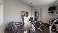 This fitness center offers a comprehensive workout environment designed to support a range of exercise routines. The space is finished with neutral gray walls and durable rubber flooring, providing a clean, functional aesthetic well-suited to an active setting. Recessed lighting throughout ensures consistent illumination, while multiple windows fitted with white horizontal blinds allow natural light to filter in, contributing to an open and energizing atmosphere. A large interior window on one wall further enhances the sense of space and connectivity within the room.

The cardio section is well-appointed, featuring a Spirit treadmill, a recumbent bike, an upright stationary bike, and an elliptical trainer, offering variety for cardiovascular workouts. A dedicated strength training area includes a multi-function cable and weight machine positioned within a recessed alcove, along with a weight bench and a rack of dumbbells for free weight exercises. A wall-mounted flat-screen television is positioned for convenient viewing during workouts. The thoughtful layout ensures adequate spacing between equipment, allowing for comfortable movement throughout the space.