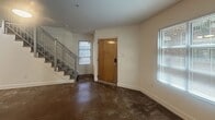 Located at 1700 E Marshall St in Richmond, Virginia, this two-floor unit offers a distinctive loft-inspired layout spread across two levels. The main floor welcomes you through a solid wood entry door into an open living area with clean white walls, wide baseboards, and a noteworthy exposed brick and timber column that adds a touch of industrial character. Natural light filters in through multiple windows dressed with horizontal blinds, brightening the space throughout. A staircase with a modern metal railing leads to the upper level, adding visual interest to the entry area. The kitchen is well-appointed with light maple cabinetry, dark granite countertops, a bold black tile backsplash, and stainless steel appliances including a refrigerator, range, and over-the-range microwave. A closet with louvered doors provides convenient storage nearby. Upstairs, the upper level is a standout feature of the unit, boasting dramatic exposed wood beam ceilings with dark-stained planking that lend a warm, rustic ambiance. Plush carpeting, a ceiling fan, and a window that brings in natural light make this level a comfortable and inviting retreat. The open railing at the stairwell maintains a sense of openness between floors, enhancing the airy, loft-like feel of the overall space.
