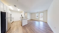 This single-floor residence offers three bedrooms, one bathroom, and a thoughtfully designed layout across a comfortable footprint. The open-concept living and kitchen area serves as the heart of the unit, featuring light-toned hardwood-style flooring that flows throughout the main spaces. Large windows and a glass exterior door flood the living area with natural light and provide outdoor views, creating a bright and welcoming ambiance.

The kitchen is well-appointed with white shaker-style cabinetry featuring brushed gold hardware, granite-patterned countertops, and a full suite of stainless steel appliances, including a range, over-the-range microwave, dishwasher, and refrigerator. A peninsula provides additional counter space and casual seating potential.

The three bedrooms are finished with plush carpet and receive natural light through window treatments. Two closets serve the unit, including a walk-in closet with wire shelving for organized storage. The bathroom features a white vanity with an undermount sink.

Connecting the rooms are two hallways that ensure a logical and functional flow throughout the residence. The overall palette of crisp white walls and neutral flooring gives the space a clean, modern feel.