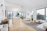 This single-floor residence is designed with a cohesive minimalist aesthetic, combining clean white walls, wide-plank light hardwood flooring, and floor-to-ceiling windows that frame sweeping views of both the waterfront and city skyline. The open-concept living and dining area is anchored by generous windows on multiple sides, bathing the space in natural light and providing an ever-present connection to the urban and aquatic landscape beyond.

The living room offers a serene, neutral palette with a plush sectional sofa, a round rattan coffee table, and a large gold-framed oval mirror that reflects the water views. Adjacent, the dining area features cane-back chairs around a natural wood table, positioned to take full advantage of the skyline panorama.

The kitchen is thoughtfully designed with flat-front, light wood cabinetry extending to the ceiling, integrated appliances, a sleek induction cooktop, and a substantial waterfall-edge island with bar seating. Bold abstract artwork punctuates the transition between kitchen and living spaces.

The primary bathroom is spa-like, featuring large-format neutral stone tiles, a freestanding soaking tub, a glass-enclosed shower, and a floating wood vanity. Bedrooms are bright and airy, each benefiting from expansive city or water views. A dedicated study nook with waterfront views and a well-connected hallway complete the thoughtfully arranged layout.