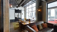 This single-floor loft space makes a striking impression with its industrial aesthetic and thoughtfully curated design. Soaring ceilings showcase exposed metal ductwork and structural elements, lending an authentic urban warehouse character to the interior. A dramatic floor-to-ceiling grid-pane window dominates the living area, flooding the space with abundant natural light and offering expansive views of the surrounding cityscape.

The living area is anchored by a rich, dark leather sofa accented with colorful throw pillows, paired with a sturdy reclaimed wood coffee table that reinforces the space's raw, tactile aesthetic. Vintage-style task lamps in a warm brass finish provide additional ambient lighting, complementing the Edison-style pendant lights that hang from the high ceiling.

Large, black-framed industrial sliding doors or partitions add a bold architectural detail, while a weathered wood accent panel introduces a rustic counterpoint to the metallic elements throughout. Dark hardwood flooring and a muted olive area rug ground the space, adding warmth underfoot. The overall ambiance strikes a compelling balance between rugged industrial design and comfortable, livable character — making it a distinctive and memorable space.