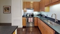 Located at 86 Fawcett Street in Cambridge, Massachusetts, this single-floor residence offers a thoughtfully designed layout that balances modern finishes with comfortable living. The kitchen is a standout feature, showcasing warm honey-toned wood cabinetry paired with dark granite countertops, stainless steel appliances including a range and refrigerator, and rich hardwood flooring. A central island provides additional prep and dining space, while under-cabinet lighting adds a polished touch throughout. The living area is bright and welcoming, featuring hardwood floors, soft blue-gray walls, and generous windows dressed with elegant drape panels and blinds that allow for flexible natural light control. The primary bedroom is distinguished by a bold terracotta accent wall, plush carpeting, and a large window that brings in ample daylight. A second bedroom offers a serene, neutral palette with soft carpet and natural light from a well-positioned window. The bathroom is clean and contemporary, with large-format tile flooring, a dark espresso vanity topped with a light countertop, and a framed mirror flanked by wall-mounted lighting. Together, these spaces create a cohesive, modern living environment well-suited to Cambridge's vibrant urban setting.