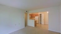 This single-floor residential unit offers a clean, open-concept layout designed for comfortable everyday living. The spacious living area is finished with wall-to-wall carpeting in a neutral tone, providing a warm and inviting atmosphere. Large, light-colored walls reflect ample ambient light throughout, giving the space an airy, open feel.

The kitchen is positioned at the heart of the unit, separated from the living area by a convenient breakfast bar that serves as both a functional workspace and a casual dining spot. Warm honey-toned wood cabinetry lines the kitchen walls, offering generous storage space, and is complemented by a white refrigerator and microwave. The kitchen floor transitions to tile, practical for a food preparation area.

Adjacent to the kitchen, a built-in shelving unit provides additional open storage, adding both utility and character to the space. A white door nearby leads to further areas of the unit. Ceiling vents are visible, indicating a central HVAC system for year-round climate control. The overall design is straightforward and functional, with neutral finishes throughout that offer a versatile backdrop easily personalized to suit any style.