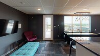 This single-floor amenity lounge is thoughtfully designed to provide a versatile social and recreational space. The kitchenette is a standout feature, showcasing rich dark wood cabinetry paired with deep granite countertops and a metallic tile backsplash. A generous bar-height counter with seating accommodates casual dining and gathering, while pendant lights with Edison bulbs hang above, adding a warm, industrial-chic aesthetic. The kitchenette is equipped with a sink and countertop appliances, making it well-suited for entertaining.

The main lounge area is defined by bold charcoal accent walls and durable patterned carpet tile flooring, creating a modern, energetic atmosphere. A large wall-mounted flat-screen television anchors one end of the room, complemented by tufted teal ottoman seating and a vibrant accent chair in deep magenta. A shuffleboard table serves as a central recreational feature, reinforcing the space's social purpose.

Natural light flows in through large windows and a full-glass exterior door that opens directly onto an outdoor terrace, where additional seating and a grill are visible. The transition between the indoor lounge and the terrace enhances the overall sense of openness. Drop ceiling panels with recessed and pendant lighting are consistent throughout, tying the various zones together into a cohesive, amenity-rich environment.