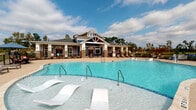 Located at 7111 Rock Fish Ln in Wilmington, NC, this single-story community amenity facility showcases a thoughtfully designed outdoor recreational complex. The centerpiece is a generously sized resort-style pool with crystal-clear water, in-pool lounge chairs, and multiple entry points via stainless steel handrails, framed by a broad, textured concrete pool deck furnished with chaise lounges for sunbathing.

The craftsman-inspired clubhouse building features brick columns, blue-gray lap siding, a gabled roofline with decorative truss detailing, and large glass-panel doors that open to the pool area. Brick columns carry through to the expansive covered pavilion areas, which provide shaded outdoor living spaces on multiple sides of the facility.

These covered areas are well-appointed with comfortable seating, including cushioned lounge chairs and sofas in blue upholstery, dining tables with woven chairs, and a wall-mounted television for outdoor entertainment. A ping-pong table adds a recreational element to one covered section. Open-air seating with umbrella-shaded bar-height tables extends poolside.

The rear covered patio offers sweeping views of lush greenery and natural surroundings beyond a black iron railing, creating a serene, nature-connected atmosphere throughout the space.