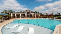 Located at 7111 Rock Fish Ln in Wilmington, NC, this single-story community amenity facility showcases a thoughtfully designed outdoor recreational complex. The centerpiece is a generously sized resort-style pool with crystal-clear water, in-pool lounge chairs, and multiple entry points via stainless steel handrails, framed by a broad, textured concrete pool deck furnished with chaise lounges for sunbathing.

The craftsman-inspired clubhouse building features brick columns, blue-gray lap siding, a gabled roofline with decorative truss detailing, and large glass-panel doors that open to the pool area. Brick columns carry through to the expansive covered pavilion areas, which provide shaded outdoor living spaces on multiple sides of the facility.

These covered areas are well-appointed with comfortable seating, including cushioned lounge chairs and sofas in blue upholstery, dining tables with woven chairs, and a wall-mounted television for outdoor entertainment. A ping-pong table adds a recreational element to one covered section. Open-air seating with umbrella-shaded bar-height tables extends poolside.

The rear covered patio offers sweeping views of lush greenery and natural surroundings beyond a black iron railing, creating a serene, nature-connected atmosphere throughout the space.