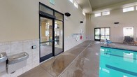 Located at 3594 N Cederblom St in Coeur d'Alene, Idaho, this single-story indoor pool facility offers a well-appointed aquatic environment designed for both recreation and relaxation. The space centers around a full-sized swimming pool with a generously wide concrete pool deck, finished with a textured, slip-resistant surface. Tile wainscoting lines the lower portion of the walls throughout, contributing to a clean and durable aesthetic suited for a wet environment. The facility features notably high ceilings that lend an open, airy feel to the interior, with upper clerestory windows allowing natural light to filter in from multiple directions. Safety equipment, including a life ring and emergency bag, is mounted along the wall, and posted pool rules are clearly visible. Adjacent to the pool area is a comfortable lounge zone furnished with rattan-style chaise lounges and teak side tables, positioned beside large windows that frame views of the surrounding courtyard and residential buildings. Glass double doors with key-card access connect the pool room to adjoining interior spaces, ensuring secure and convenient entry. Wall-mounted speakers suggest the space is equipped for ambient audio, enhancing the overall atmosphere of this well-maintained community aquatic facility.
