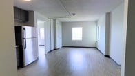 This single-floor space offers a clean, functional layout designed for comfortable everyday living. The main living area is spacious and open, finished with light-toned hardwood-style flooring that reflects natural light and lends a bright, airy feel to the interior. Two windows along the exterior wall allow generous daylight to fill the room, creating a welcoming atmosphere throughout the day. The walls are painted in a neutral gray tone, complemented by dark baseboards that provide a subtle contrast and a polished, modern look. A textured ceiling adds a classic touch to the overall aesthetic. The kitchen area is positioned adjacent to the living space and is equipped with a full-size refrigerator, making it practical for daily use. Closet storage is thoughtfully integrated near the interior corridor, offering ample organizational space. A hallway visible from the main room suggests additional rooms or utility spaces beyond, contributing to the unit's overall functionality. The space maintains a neutral, move-in-ready palette throughout, making it highly adaptable to a variety of furnishing styles and personal preferences. Overall, the layout balances openness with practicality in a well-maintained, single-level setting.