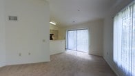 This single-floor space offers an open and airy layout designed for comfortable living. The main living area is finished with neutral-toned carpet and bright white walls, creating a clean and inviting atmosphere. A large sliding glass door fitted with vertical blinds serves as a focal point, allowing an abundance of natural light to pour into the room while providing access to an outdoor area with visible greenery beyond.

Adjacent to the living space is a kitchen area, partially separated by a half-wall pass-through. The kitchen features light-colored cabinetry and is equipped with under-cabinet fluorescent lighting, lending a functional and practical feel. The pass-through counter enhances the open-concept flow between the kitchen and living areas, making the space feel connected and social.

The ceilings are a standard height with recessed vents, and the overall color palette—comprising whites and soft neutrals—contributes to a light and spacious feel throughout. The minimalist design of the space allows for flexible furniture arrangements, making it adaptable to a variety of lifestyle needs.