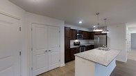 This single-floor residence offers a thoughtfully designed layout centered around a standout kitchen that blends style and functionality. The kitchen features rich, dark wood cabinetry extending to the upper walls, providing generous storage throughout. Light granite countertops offer a striking contrast, complementing the full suite of stainless steel appliances, which includes a range with electric cooktop, built-in microwave, and a French door refrigerator. A kitchen island anchors the space, topped with matching granite and illuminated by two industrial-style pendant lights that add a modern, polished touch. Recessed lighting throughout ensures the kitchen is bright and well-lit. Light-toned hardwood flooring runs through the space, adding warmth and cohesion to the overall aesthetic. A set of classic paneled double doors, finished in crisp white, provides access to additional storage or a utility area, adding to the practicality of the layout. The open and efficient design of this residence makes it well-suited for everyday living, with clean lines, quality finishes, and a neutral palette that creates a welcoming and functional atmosphere throughout.