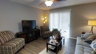 This single-floor residence offers a thoughtfully arranged living space designed for both comfort and functionality. The living room serves as the heart of the home, featuring smooth wood-style flooring that adds warmth and continuity throughout the space. A ceiling fan with an integrated light fixture is centrally mounted, providing both air circulation and ambient lighting. Large sliding glass doors dressed with vertical blinds allow generous natural light to fill the room while offering access to the outdoors. The living area is well-proportioned, comfortably accommodating a seating arrangement with striped accent chairs, a dark wood entertainment console, and a classic coffee table that anchors the space. Warm accent lighting from a floor lamp enhances the inviting atmosphere during evening hours. Neutral gray walls provide a versatile backdrop that complements the rich, dark wood furnishings and earthy tones present throughout. The overall ambiance is relaxed and residential, with a practical layout that maximizes the use of available space. The combination of natural light, warm tones, and functional furnishings makes this a comfortable and livable single-floor home suited for everyday living.