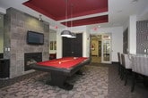 This single-floor amenity space is thoughtfully designed to accommodate a variety of social and leisure activities within a cohesive, modern interior. The layout is organized into distinct functional zones, each contributing to the overall inviting atmosphere.

A standout feature is the game area, anchored by a full-size billiards table in a rich red felt finish, positioned beneath a bold coffered ceiling with deep crimson accents and a statement pendant light. Adjacent to the table, a floor-to-ceiling stone-clad fireplace with a wall-mounted television creates a warm, lounge-like focal point.

A sunlit seating alcove lines an expansive floor-to-ceiling wraparound window wall, where clusters of patterned red accent chairs and slim side tables invite conversation while offering views of surrounding greenery. The patterned carpet throughout the space ties the zones together visually.

A secondary lounge area features a tufted sofa, patterned armchairs, and French doors that open directly onto a covered outdoor balcony, providing seamless indoor-outdoor connectivity and natural light. A glimpse of a dedicated workspace is also visible, offering a quieter retreat within the broader space.