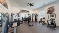 Located at 900 SE Third Ave in Ocala, Florida, this dedicated fitness center offers a thoughtfully organized workout facility on a single floor. The space is designed to maximize functionality, with durable rubber-speckled flooring providing both safety and comfort underfoot throughout the entire area.

The room is outfitted with a diverse selection of exercise equipment catering to both strength training and cardiovascular fitness. Strength equipment includes a multi-function cable machine, a leg press machine, a dedicated dumbbell rack with a full range of free weights, and a weight bench, while cardio options feature a treadmill, elliptical trainer, recumbent bike, and aerobic step platform. Large wall-mounted mirrors along one side of the room enhance the sense of space and allow users to monitor their form during workouts.

Natural light filters in through multiple windows fitted with white blinds, keeping the space bright and inviting. Recessed lighting and a ceiling fan with overhead lighting ensure the room remains well-lit and comfortable year-round. A wall-mounted flat-screen TV provides entertainment, and a coat rack with a storage bench near the entrance adds practical convenience. The white walls are accented with vibrant equestrian-themed artwork, lending the space a distinctive, local character reflective of Ocala's renowned horse country heritage.