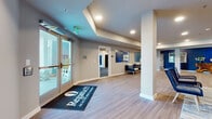 This single-story clubhouse for Regatta 55, an active adult community, presents a thoughtfully designed gathering space with a cohesive nautical-inspired aesthetic throughout. Entry is made through a set of full-glass double doors framed by industrial-style wall sconces, opening into a bright lobby with light wood-look flooring and a branded welcome mat.

The open-plan interior is anchored by bold navy blue accent walls that contrast with neutral gray tones, recessed lighting, and white structural columns. A spacious lounge area features a comfortable arrangement of sofas, leather chairs, and accent seating atop patterned area rugs, creating an inviting communal gathering space. Framed wall art and decorative sculptures reinforce the nautical design theme throughout.

A dedicated leasing and administrative office area is carpeted in a deep blue tone and includes a workstation desk, seating for guests, a circular meeting table, and a pass-through window connecting to adjacent spaces. A separate consultation or meeting room with a glass-panel door provides additional privacy.

The layout also includes a dining area visible in the background, complete with dark furnishings, and a flex space that can accommodate community activities. Natural light filters in through multiple windows, complementing the warm recessed lighting that illuminates the entire facility.