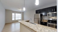 This single-floor, 2-bedroom, 2-bathroom residence offers a thoughtfully designed open-concept layout that maximizes functionality and flow. The kitchen serves as a central highlight, featuring granite countertops with subtle beige and gray veining, dark espresso cabinetry, and stainless steel appliances including a full-sized refrigerator. A kitchen island with an undermount sink and pendant lighting above creates a natural gathering point and provides additional workspace. The open layout connects the kitchen seamlessly to the living area, which benefits from a window that allows natural light to fill the space. The clean white walls and light flooring throughout contribute to a bright, contemporary ambiance. Modern recessed and pendant lighting fixtures ensure consistent illumination across the living and kitchen areas. The overall design aesthetic is sleek and minimalist, with dark cabinetry contrasting against light countertops and neutral walls. The efficient use of space throughout the unit makes it well-suited for comfortable everyday living while maintaining a polished, modern appearance.